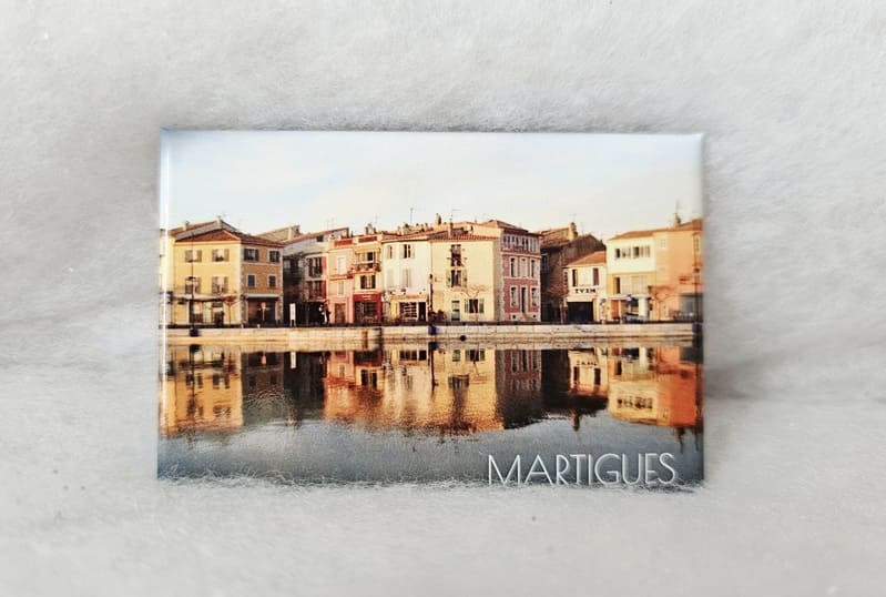 Imán con una foto del Quai des Girondins de Martigues, reconocible por sus coloridas fachadas y reflejos en el agua. Foto de un paseo marítimo en Martigues con sus canales y coloridas fachadas. Los reflejos en el agua añaden un toque romántico a la imagen. Imán a la venta en la Oficina de Turismo de Martigues