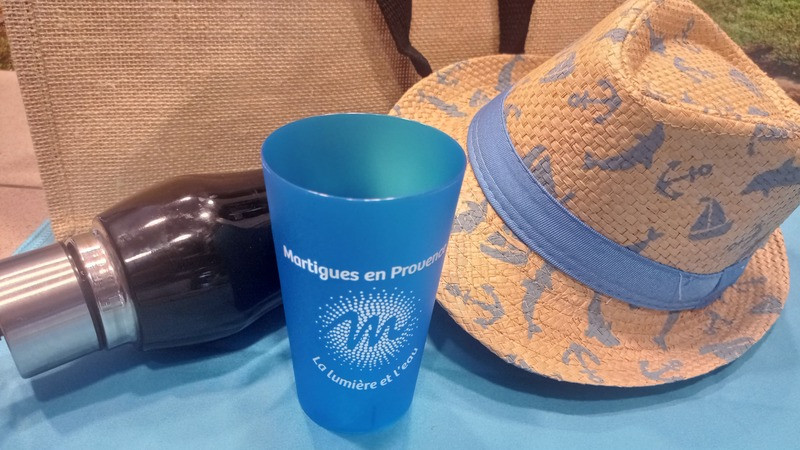 Blue cup from the Soleil range featuring the logo of the Martigues Tourist Office, placed on a blue beach towel alongside a straw hat, a beach bag, and a water bottle