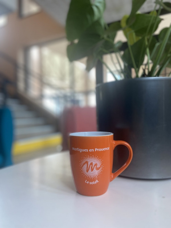 Mug orange vif élégamment posé sur une table grise, avec une plante en arrière-plan et un escalier flou qui ajoute de la profondeur à la scène