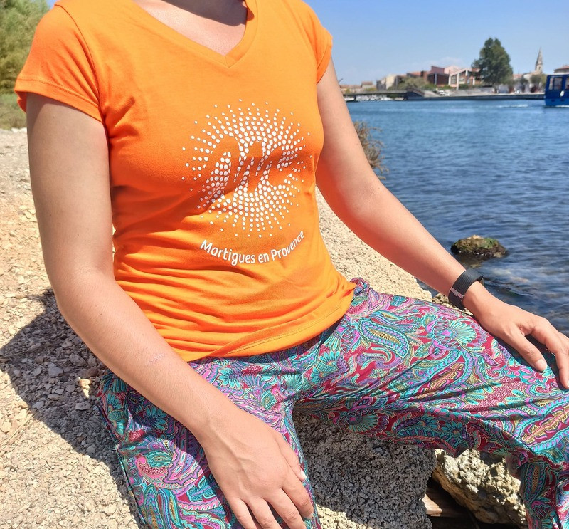 Tee-shirt orange de la Gamme Soleil, orné du logo blanc de l'Office de Tourisme au centre, porté par un modèle féminin avec vue sur le canal en arrière-plan