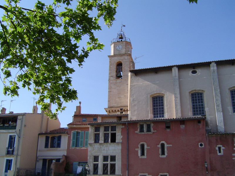 L'église Marie Madeleine et son clocher pris de côté avec le bas de la façade rouge et le haut de la façade blanche. Des maisons aux volets verts sont accolés à l'église. Un arbre laisse apercevoir ses branches au premier plan.