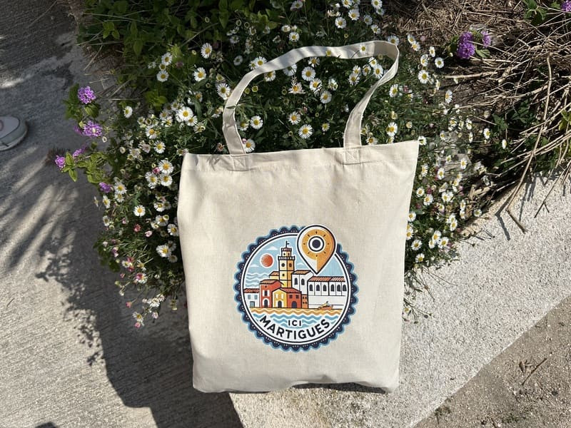 Tote bag beige avec dessin sérigraphié orange et rouge de Martigues, texte « Ici Martigues » et symbole de localisation au centre