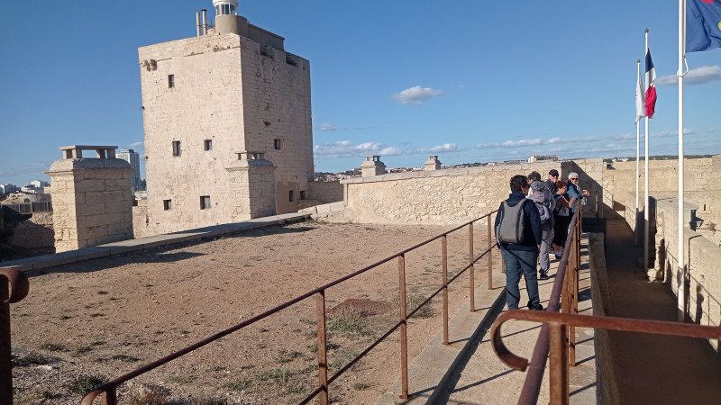 Le groupe marche sur une passerelle sur les hauteurs du château. La tour du Fort apparaît à droite, on ne voit pas le bas du bâtiment. Un drapeau français flotte dans les airs, accroché à un mât. 
