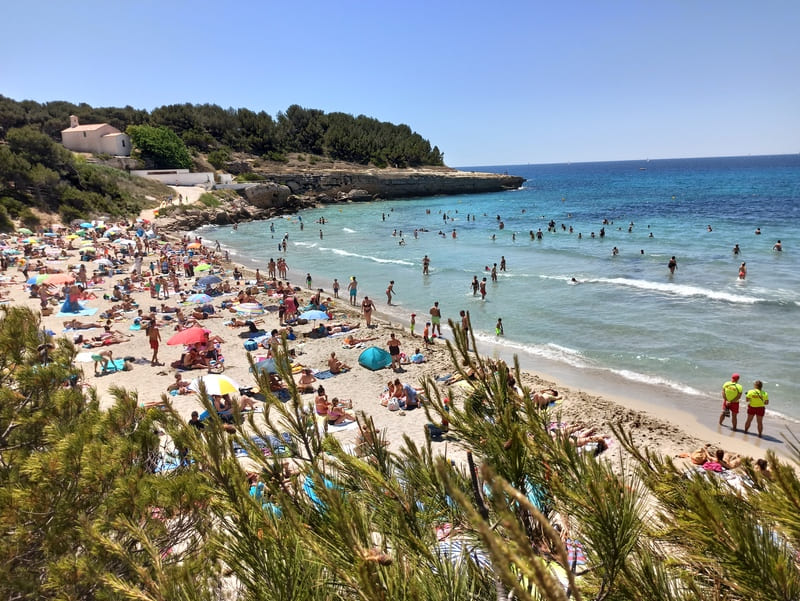 Photo prise sur les hauteurs de la plage de Sainte-Croix qui forme une anse abritée par les falaises environnantes. L'eau a un crescendo de bleu turquoise du Sud de la France. Des touristes bronzent sur la plage de sable, la chapelle est visible en face - © Otmartigues / KarimK