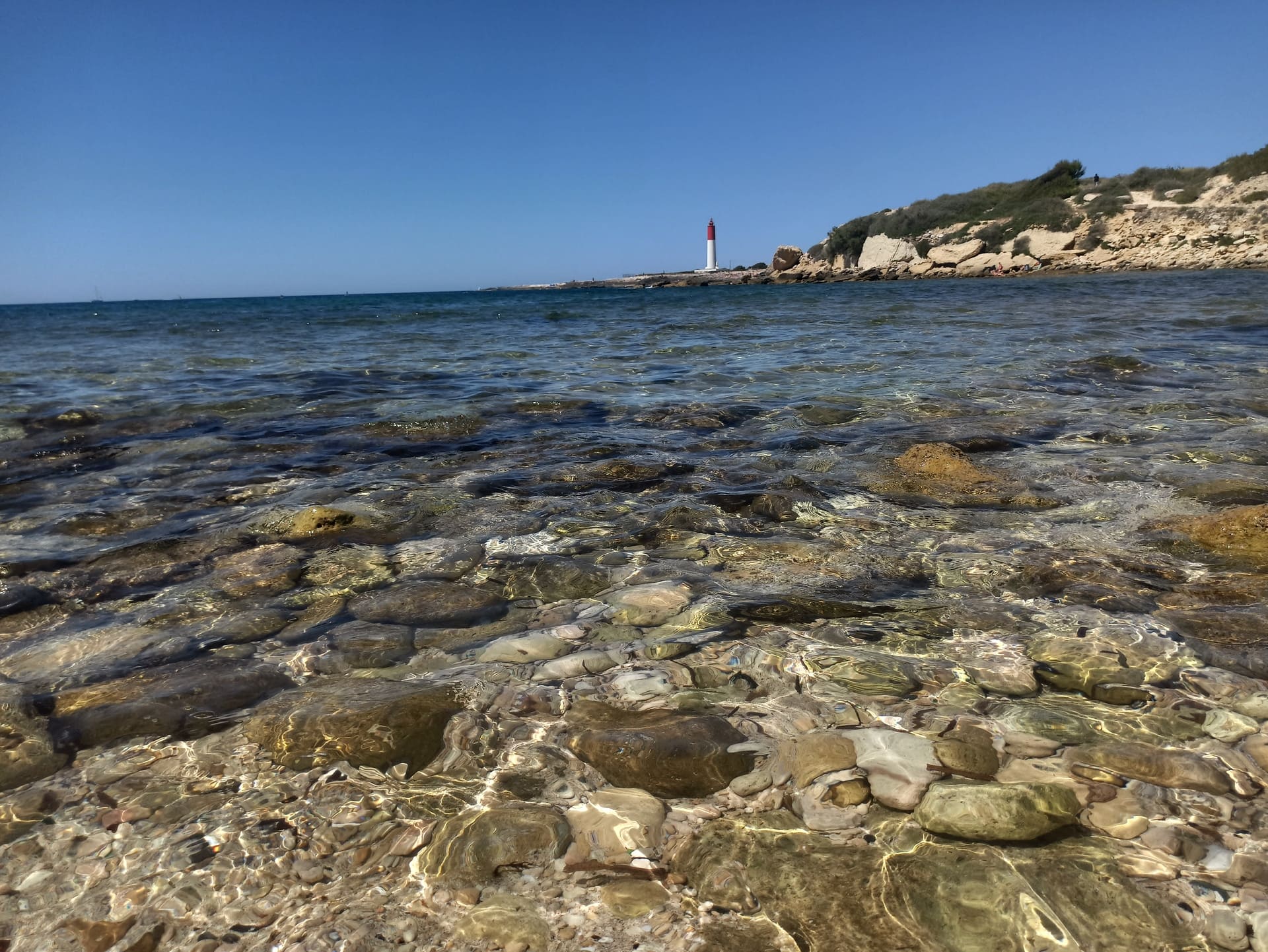 Les pieds dans l'eau avec un fond clair qui laisse apercevoir le sol rocheux. Au loin, le phare de La Couronne se dresse fièrement parmi les collines aux sommets verdoyants - © Otmartigues / KarimK