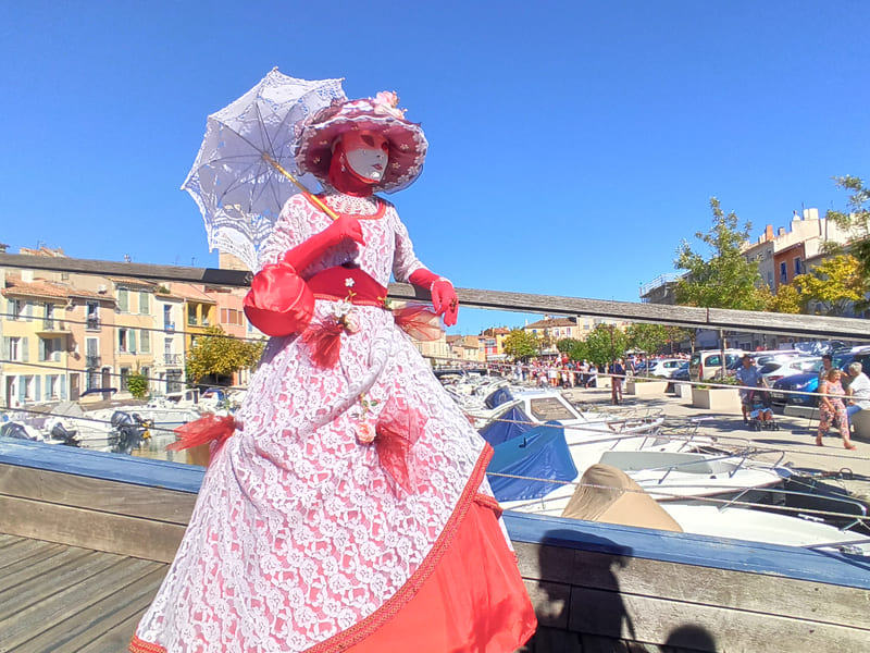 Vestido rojo de capas con detalles provenzales, sombrilla y bolso de señora, con el rostro completamente enmascarado, como en Venecia, durante el desfile de Martigues. - © Otmartigues / KarimK