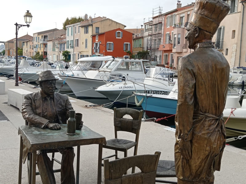 Vue prolongée des statues de Bourvil et Fernandel représentant la scène au restaurant dans le film La Cuisine au Beurre. Des sièges sont libres pour une photo souvenir devant les façades colorées - © Otmartigues / MyriamF